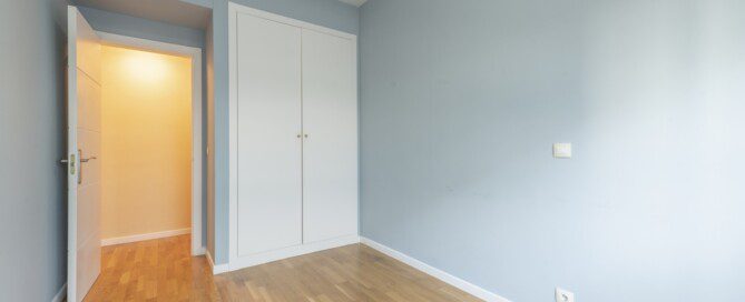 Bedroom of an empty house with laminated oak flooring with a white built-in wardrobe, ducted air conditioning and white carpentry.